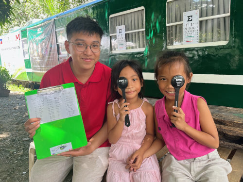 Canadian Vision Care - Philippines Eye Train Image of two children, each holding ocluders in front of one eye, next to an optometrist. Behind them is the Eye Train, a train car that was converted into an eye clinic.