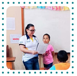 Photo of a teacher in Guatemala beside a student, holding a book which the student is reading.