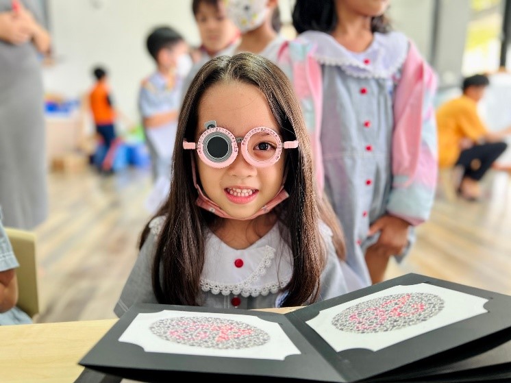 A photo of a young girl wearing trial lens glasses and smiling, in front of books with eye exam images.